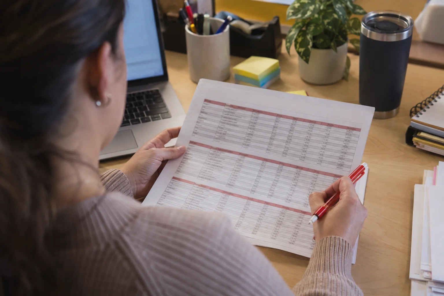 Merchandising assistant reviewing a printed promotion price list with a red pen, a laptop open beside her