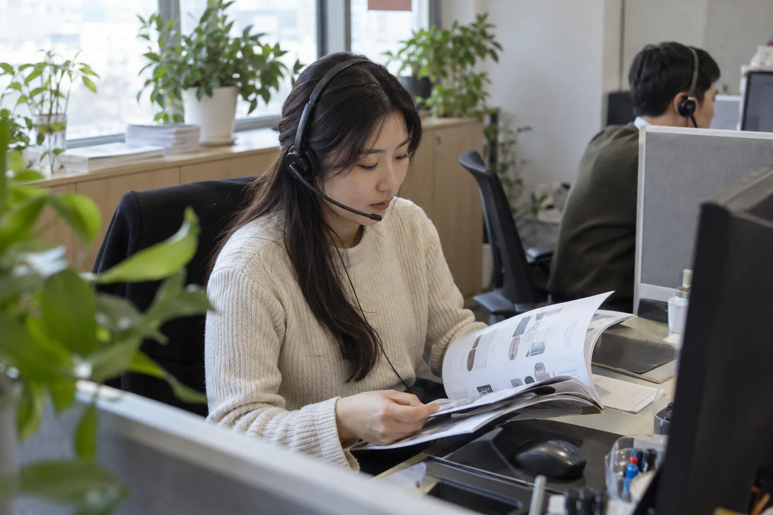 작은 상담 코너에서 헤드셋을 쓴 크림색 스웨터 차림의 상담원이 제품 사양서 책자를 넘기는 모습
