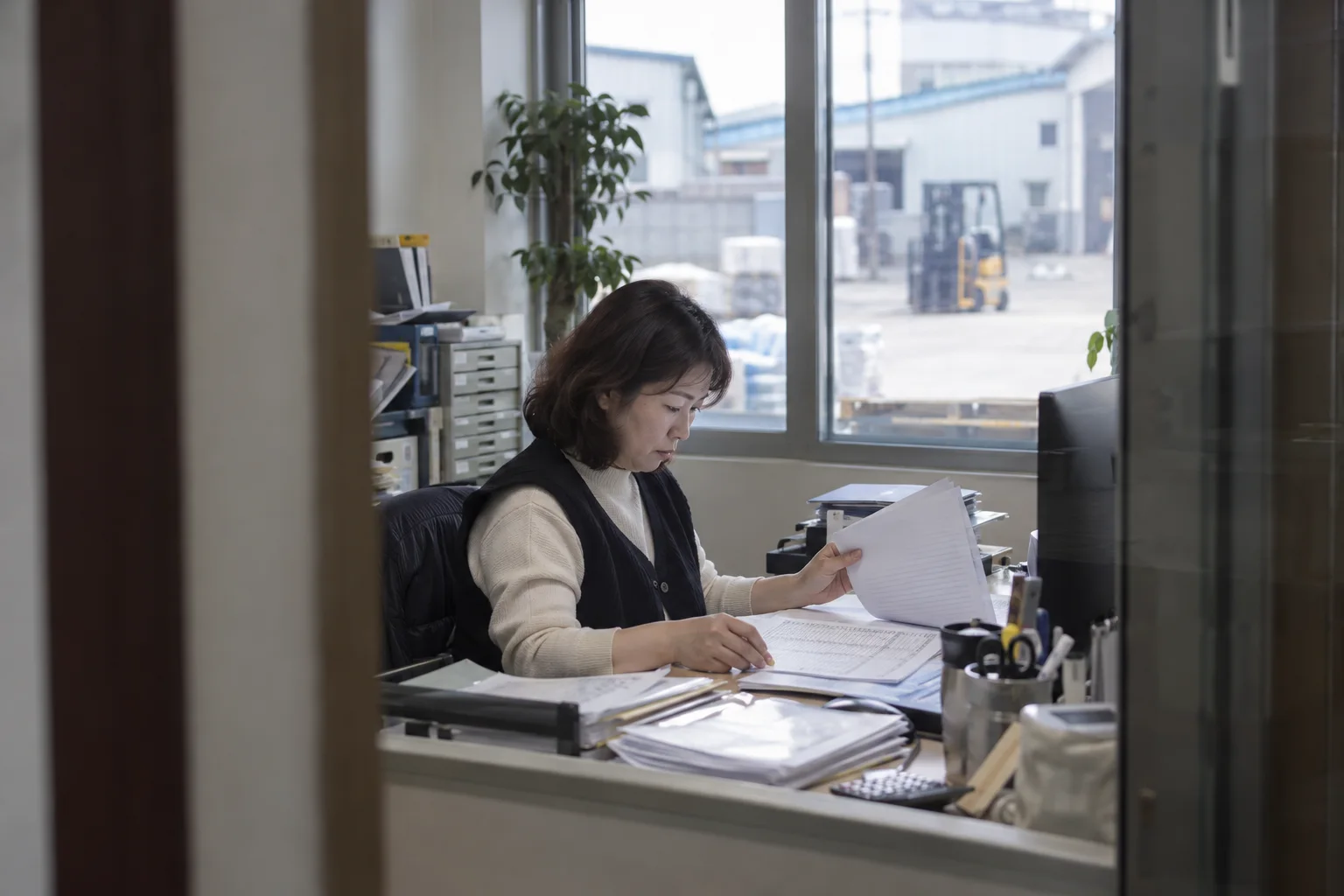 공장 사무실 유리문 너머로 관리 담당 직원이 급여 대장 출력물을 넘겨 보고 창밖으로 지게차가 선 마당이 보이는 모습